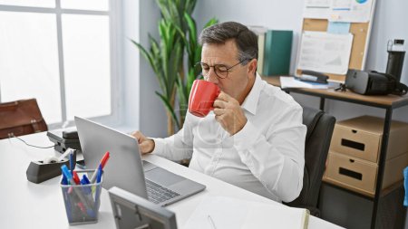 Photo for Middle-aged man in glasses sipping coffee while working on laptop in bright office. - Royalty Free Image