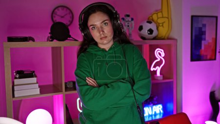 A confident young woman in a green hoodie with crossed arms stands in a colorful gaming room at night.
