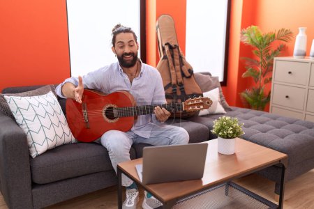 Foto de Hombre hispano con barba tocando la guitarra clásica en casa celebrando el logro con sonrisa feliz y expresión ganadora con la mano levantada - Imagen libre de derechos