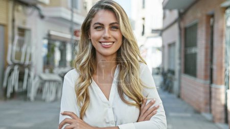 Photo for Cheerful, confident young blonde woman enjoying the sunny city atmosphere, standing on a street with a joyful smile and relaxed arms crossed gesture - Royalty Free Image