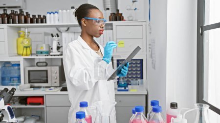 Foto de Apasionada científica afroamericana en guantes de laboratorio, fascinada por el poder de la investigación médica, profundamente absorta en tomar notas en un laboratorio de ciencias de interior - Imagen libre de derechos