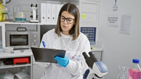 Foto de Una científica hispana enfocada examina notas en un laboratorio rodeado de equipo. - Imagen libre de derechos