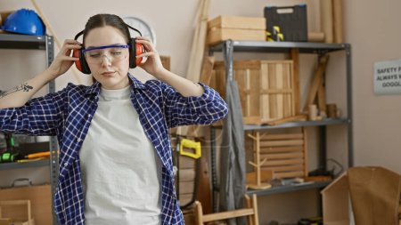 Caucasian woman wearing safety gear in a carpentry workshop surrounded by wooden furniture and tools.
