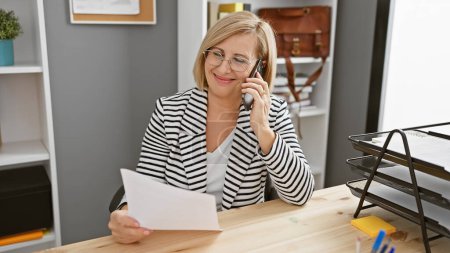 Téléchargez les photos : Une femme blonde professionnelle d'âge moyen s'est concentrée sur la paperasserie tout en conversant sur son téléphone dans un cadre de bureau moderne. - en image libre de droit