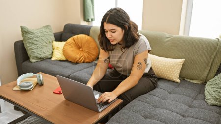 Téléchargez les photos : Une jeune femme concentrée utilise un ordinateur portable dans un salon moderne orné d'oreillers colorés. - en image libre de droit