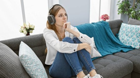 Pensive young caucasian woman wearing headphones sitting on a gray sofa in a cozy living room.