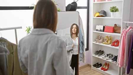 Photo for Young woman trying on jacket in a modern dressing room, with mirror and shelves of shoes and bags. - Royalty Free Image