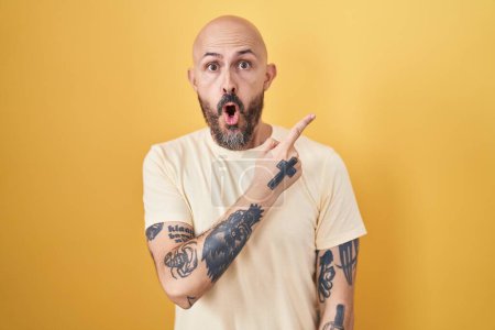 Photo for Hispanic man with tattoos standing over yellow background surprised pointing with finger to the side, open mouth amazed expression. - Royalty Free Image