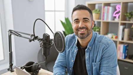 Photo for A smiling hispanic man with a beard in a studio setting with microphone and laptop, projecting a professional yet casual demeanor. - Royalty Free Image