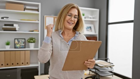 A joyful mature woman celebrates success with a clipboard in a modern office setting, indicating achievement and job satisfaction.