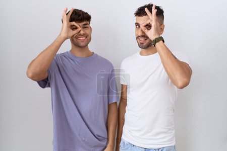 Téléchargez les photos : Homosexuel gay couple debout sur blanc fond faire ok geste avec main sourire oeil regarder à travers doigts avec heureux visage. - en image libre de droit