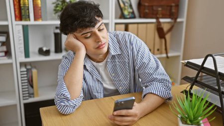 Young man bored at office desk with smartphone, showing a moment of work fatigue or break time.