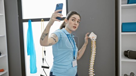 Foto de Mujer joven caucásica en clínica hospitalaria tomando selfie con modelo de columna vertebral en interiores - Imagen libre de derechos