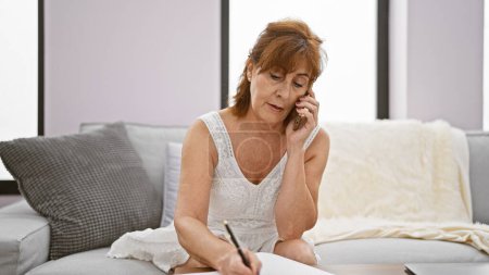 Foto de Mujer madura hablando por teléfono móvil y escribiendo notas en papel en un ambiente acogedor sala de estar. - Imagen libre de derechos