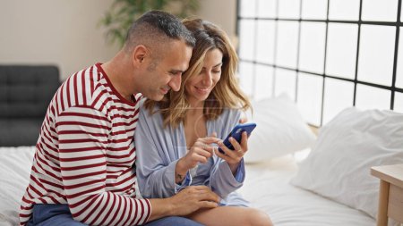 Téléchargez les photos : Homme et femme couple assis sur le lit en utilisant smartphone dans la chambre - en image libre de droit