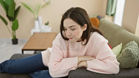 Foto de Mujer joven y relajada en un suéter rosa casual sentada en casa en un sofá acogedor, profundo en el pensamiento - Imagen libre de derechos