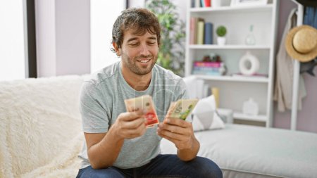 Photo for Confident young man with blond beard, smiling and sitting on sofa at home, counting a wealth of israeli shekels banknotes as his investment payoff - Royalty Free Image