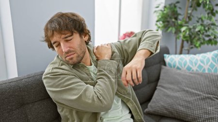 Foto de Hombre joven adulto con barba que experimenta dolor de hombro en el interior, sentado en un sofá en casa, con una expresión dolorosa. - Imagen libre de derechos