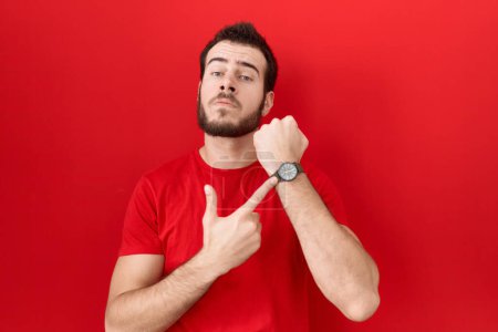 Téléchargez les photos : Jeune homme hispanique portant un t-shirt rouge décontracté à la hâte pointant vers regarder le temps, l'impatience, regardant la caméra avec une expression détendue - en image libre de droit