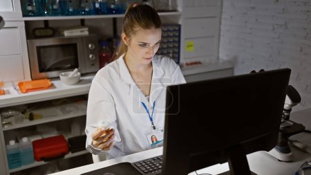 Eine fokussierte junge Frau arbeitet in einem Labor an einem Computer und hält eine Tablettenflasche in der Hand