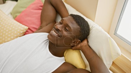 Photo for An attractive african american man relaxes smiling on colorful pillows in a bright bedroom setting. - Royalty Free Image