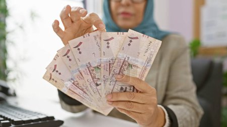 Téléchargez les photos : Une femme dans un hijab compte les factures saoudiennes à son bureau, suggérant une transaction commerciale ou une gestion financière à l'intérieur. - en image libre de droit