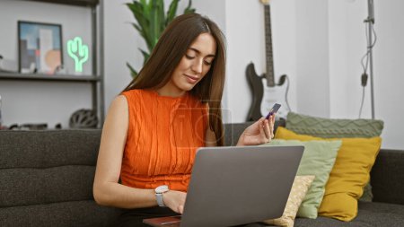 Téléchargez les photos : Une femme en orange évalue une carte de crédit tout en travaillant sur un ordinateur portable dans un intérieur de maison élégant. - en image libre de droit