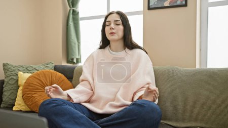 Photo for A tranquil young woman meditating on a cozy sofa in a well-lit living room, embodying mindfulness and serenity. - Royalty Free Image