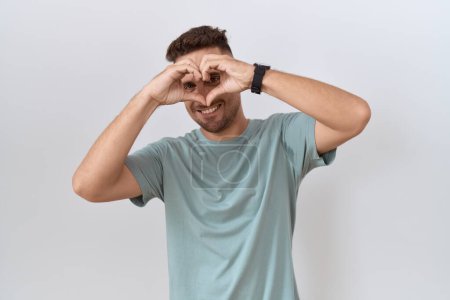 Foto de Hombre hispano con barba de pie sobre fondo blanco haciendo forma de corazón con la mano y los dedos sonriendo mirando a través de signo - Imagen libre de derechos