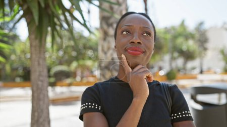 Coole, attraktive afrikanisch-amerikanische Frau, die mit zweifelndem Gesichtsausdruck auf einer sonnigen Stadtstraße steht und in entspannter, urbaner Lifestylemode über eine Idee nachdenkt