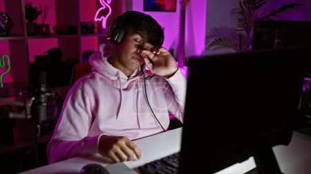 Foto de Joven streamer hispano cansado, sesión de juegos nocturnos en cuarto oscuro, luchando por seguir jugando videojuegos en la computadora - Imagen libre de derechos