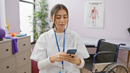 Eine junge hispanische Gesundheitshelferin benutzt ein Smartphone in einem Reha-Raum der Klinik, im Hintergrund ein Rollstuhl.