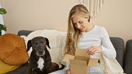 Photo for A young woman unboxing with an attentive black dog beside her on a cozy living room couch. - Royalty Free Image