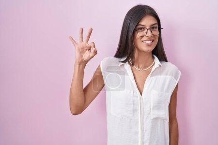 Foto de Morena joven mujer de pie sobre fondo rosa con gafas sonriendo positiva haciendo signo de ok con la mano y los dedos. expresión exitosa. - Imagen libre de derechos