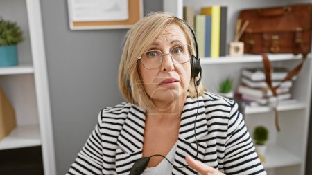 Foto de Una mujer de mediana edad perpleja con el pelo rubio usando un auricular en un entorno de oficina moderno. - Imagen libre de derechos
