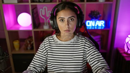 Photo for A young hispanic woman with headphones in a colorful gaming room looks surprised at night. - Royalty Free Image