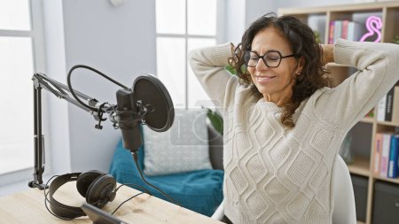 Hispanic woman relaxing in a radio studio with microphone and headphones on desk.