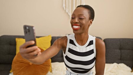 Téléchargez les photos : Joyeuse femme afro-américaine assise sur le canapé à la maison, prenant un selfie attrayant avec son téléphone dans le salon, rayonnant de positivité et de bonheur. - en image libre de droit