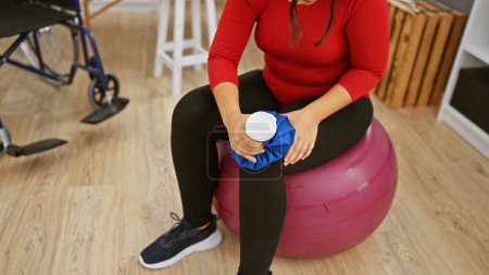 Photo for Hispanic woman exercising with ice pack and ball in rehab clinic's gym room - Royalty Free Image