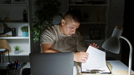 Ernsthafter junger lateinischer Mann nagelt nächtliche Büroarbeit, liest Business-Doku auf Laptop, zweifelt, aber entspannt, Managerleben drinnen