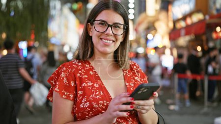 Téléchargez les photos : Joyeux belle femme hispanique dans des lunettes profitant d'une conversation de texte de nuit sur son smartphone à la célèbre rue dotonbori d'osaka - en image libre de droit