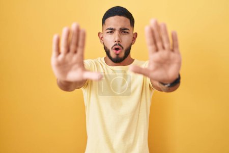 Foto de Joven hombre hispano de pie sobre fondo amarillo haciendo gesto de stop con palmas de las manos, enojado y expresión de frustración - Imagen libre de derechos