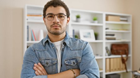Photo for Casual young man with glasses and beard standing arms crossed in a modern office setting. - Royalty Free Image