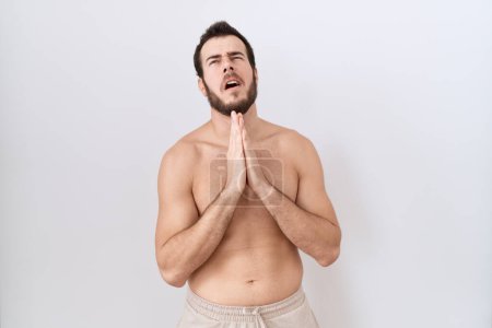 Téléchargez les photos : Jeune homme hispanique debout torse nu sur fond blanc suppliant et priant avec les mains ainsi que l'expression de l'espoir sur le visage très émotionnel et inquiet. mendier. - en image libre de droit