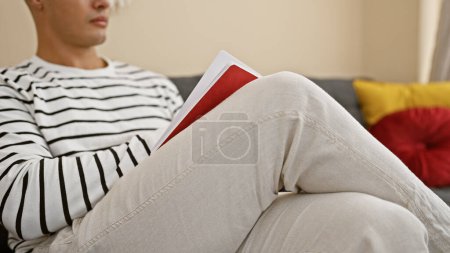 Téléchargez les photos : Jeune homme hispanique attrayant et intelligent, plongé dans un livre, confortablement assis sur un canapé à la maison. étudier tranquillement dans le salon confortable, absorber les connaissances tout en se relaxant à l'intérieur. - en image libre de droit