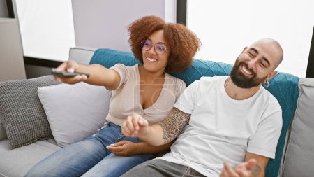 Téléchargez les photos : Joyeux et beau couple profiter du plaisir ensemble à la maison assis confortablement sur leur canapé salon, rire et changer de chaîne en toute confiance, regarder leur film préféré à la télévision - en image libre de droit