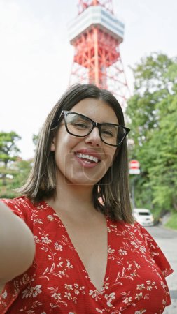 Schöne hispanische Frau mit Brille, genießt ihr Tokyo-Reiseabenteuer und macht ein fröhliches Selfie am berühmten Tokyo-Turm
