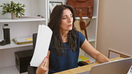 Photo for Mature hispanic woman holding documents while working thoughtfully in a modern office setting, representing diversity in the workplace. - Royalty Free Image