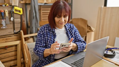 Foto de Una mujer de mediana edad usa un teléfono inteligente en su taller de carpintería mientras un portátil está sobre la mesa. - Imagen libre de derechos