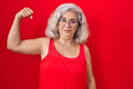 Téléchargez les photos : Femme d'âge moyen avec les cheveux gris debout sur fond rouge personne forte montrant muscle du bras, confiant et fier de la puissance - en image libre de droit